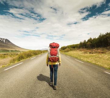 An outdoorsman with a red backpack strides down a deserted road, enveloped by majestic mountains and towering trees under a partly cloudy sky. | outdoorsurvivors.com