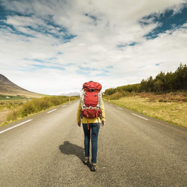 An outdoorsman with a red backpack strides down a deserted road, enveloped by majestic mountains and towering trees under a partly cloudy sky. | outdoorsurvivors.com