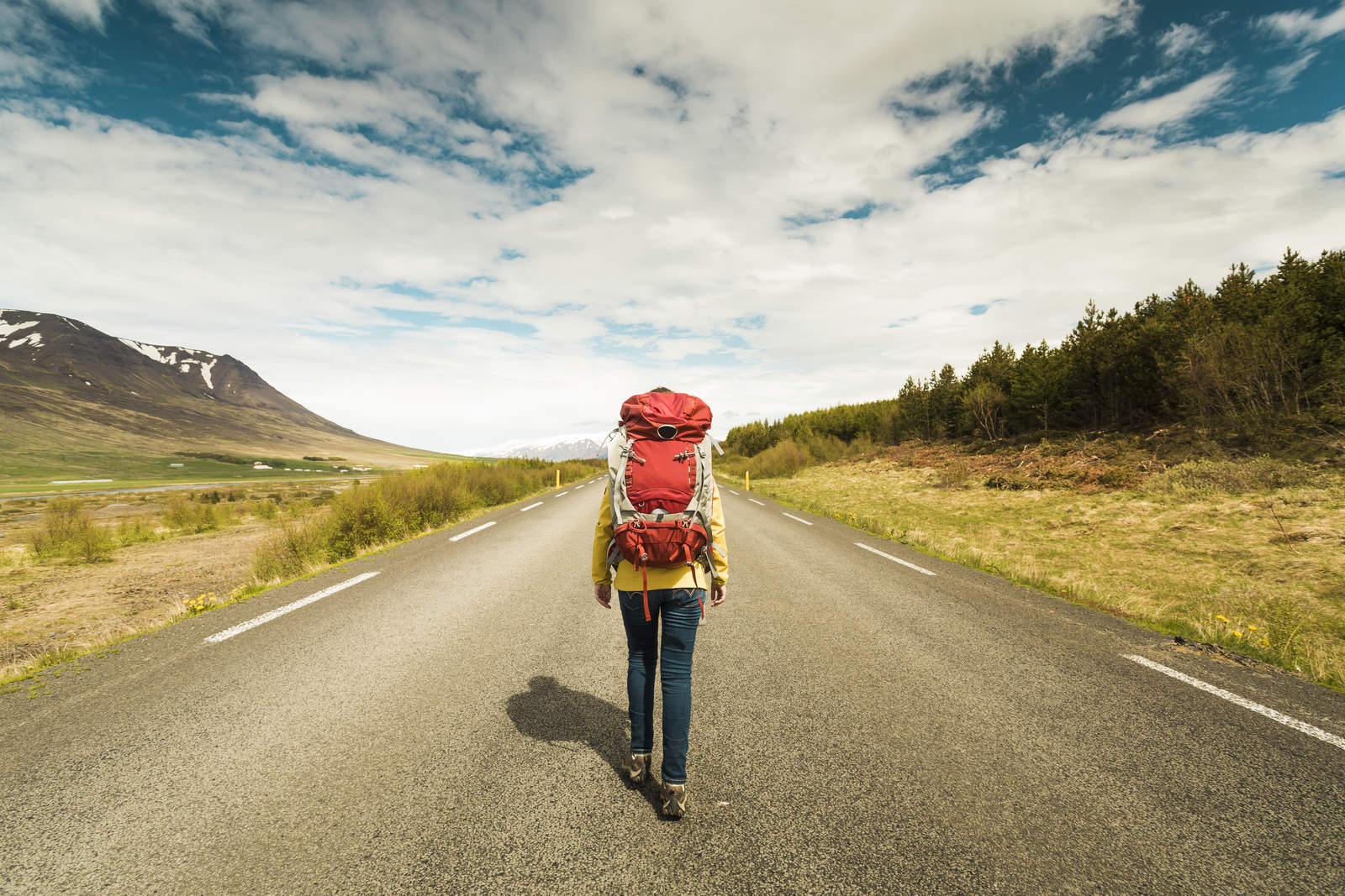 An outdoorsman with a red backpack strides down a deserted road, enveloped by majestic mountains and towering trees under a partly cloudy sky. | outdoorsurvivors.com