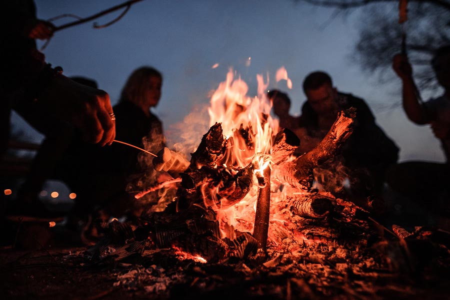 People sit around a campfire roasting marshmallows on sticks, with flames illuminating the scene against a dusk sky. | outdoorsurvivors.com People sit around a campfire roasting marshmallows on sticks, with flames illuminating the scene against a dusk sky. | outdoorsurvivors.com