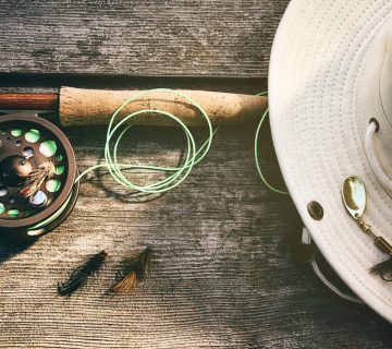 A fishing rod and reel, green line, beige hat with a lure, and three flies sit on a wooden surface, perfect for fly fishing beginners. | outdoorsurvivors.com