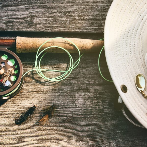 A fishing rod and reel, green line, beige hat with a lure, and three flies sit on a wooden surface, perfect for fly fishing beginners. | outdoorsurvivors.com