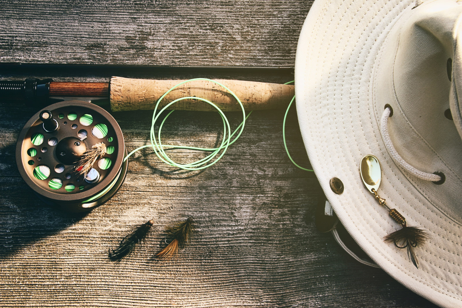A fishing rod and reel, green line, beige hat with a lure, and three flies sit on a wooden surface, perfect for fly fishing beginners. | outdoorsurvivors.com