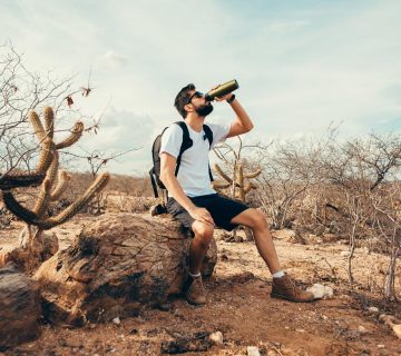A man in shorts and a t-shirt sits on a rock in a dry, desert landscape with cacti, sipping from a water bottle—enjoying a break during his desert hiking adventure. | outdoorsurvivors.com