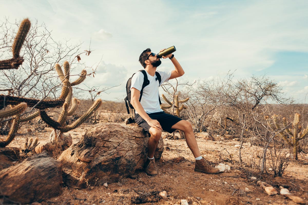 A man in shorts and a t-shirt sits on a rock in a dry, desert landscape with cacti, sipping from a water bottle—enjoying a break during his desert hiking adventure. | outdoorsurvivors.com