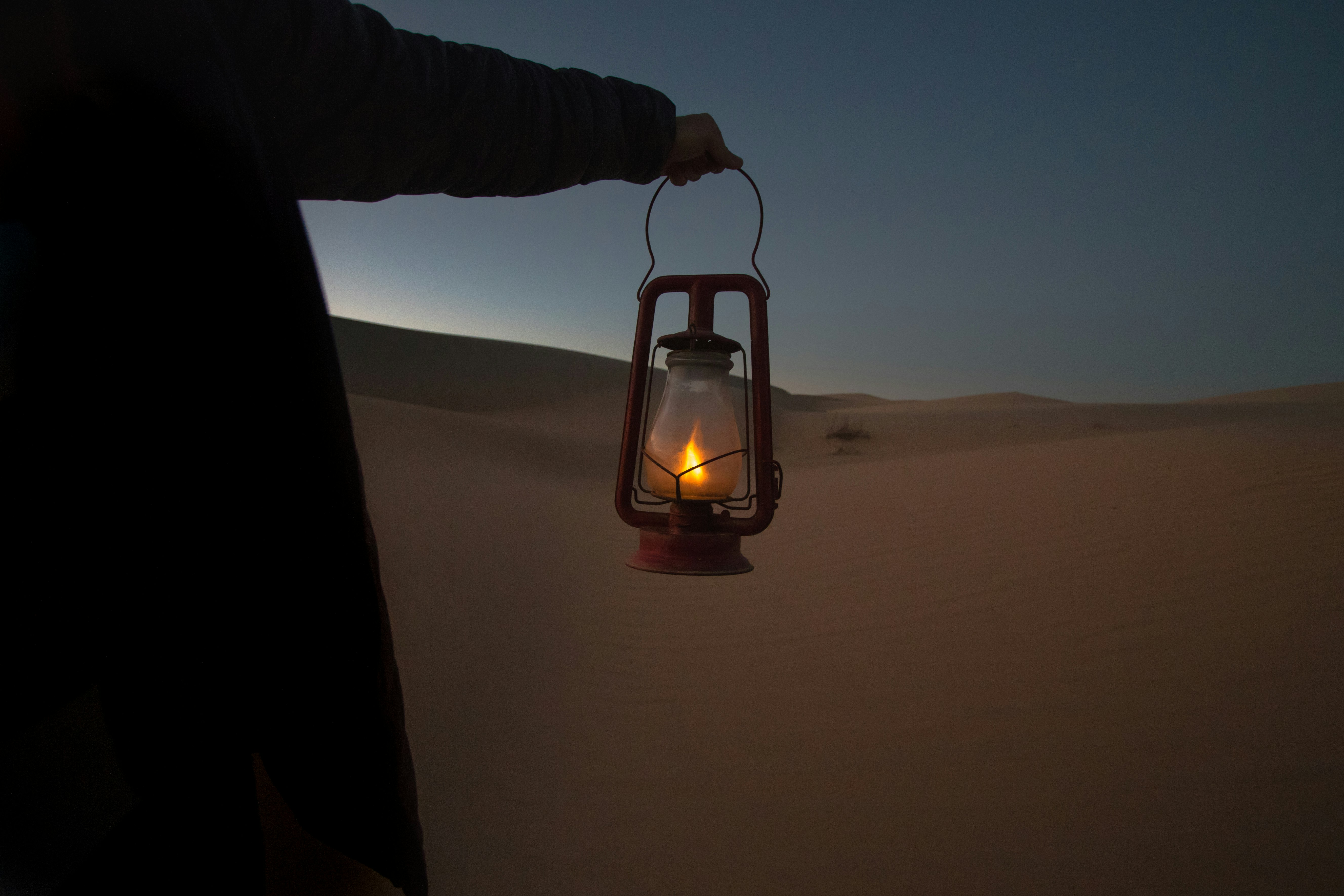A person holds a lit lantern in a sandy desert at dusk, with silhouetted dunes and a darkening sky in the background | outdoorsurvivors com