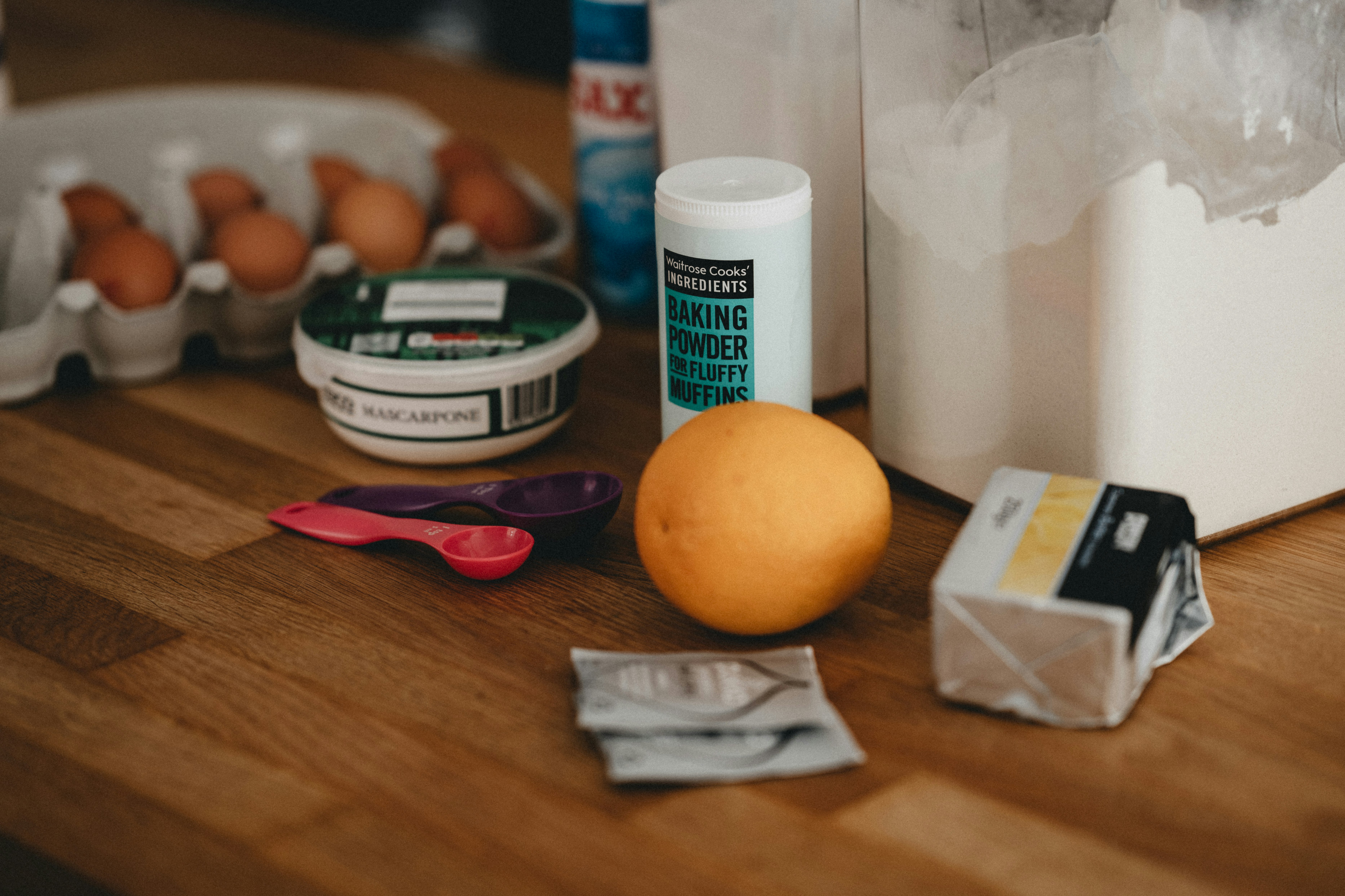Eggs, mascarpone, baking powder, a lemon, butter, measuring spoons, and packets on a wooden countertop, with a white container and cleaning spray in the background | outdoorsurvivors com