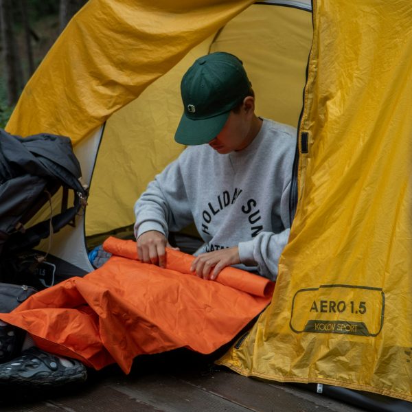 Person sitting at the entrance of a yellow tent, folding an orange sleeping bag, with a large backpack and hiking boots nearby in a forest setting. | outdoorsurvivors.com Person sitting at the entrance of a yellow tent, folding an orange sleeping bag, with a large backpack and hiking boots nearby in a forest setting | outdoorsurvivors com
