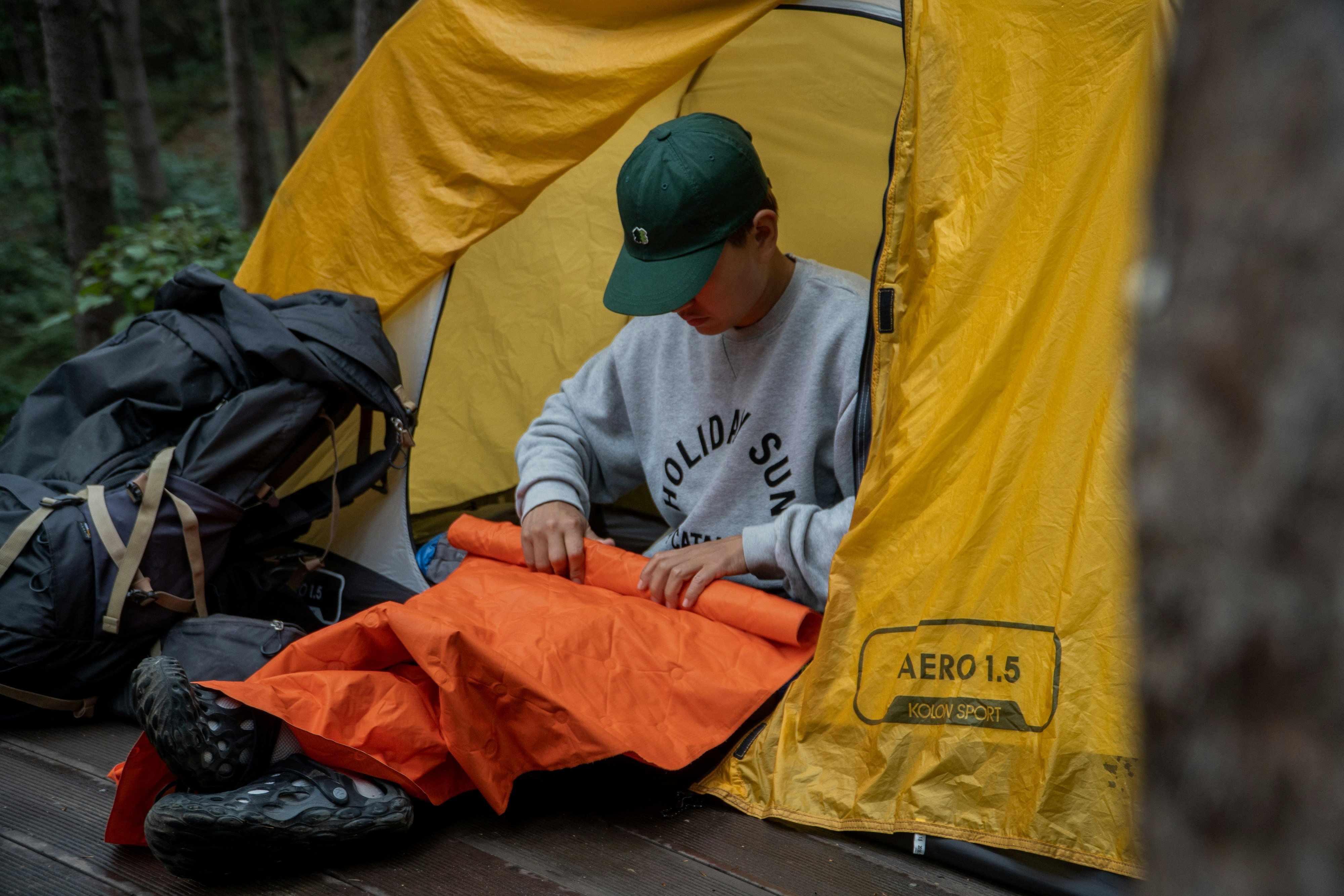 Person sitting at the entrance of a yellow tent, folding an orange sleeping bag, with a large backpack and hiking boots nearby in a forest setting | outdoorsurvivors com