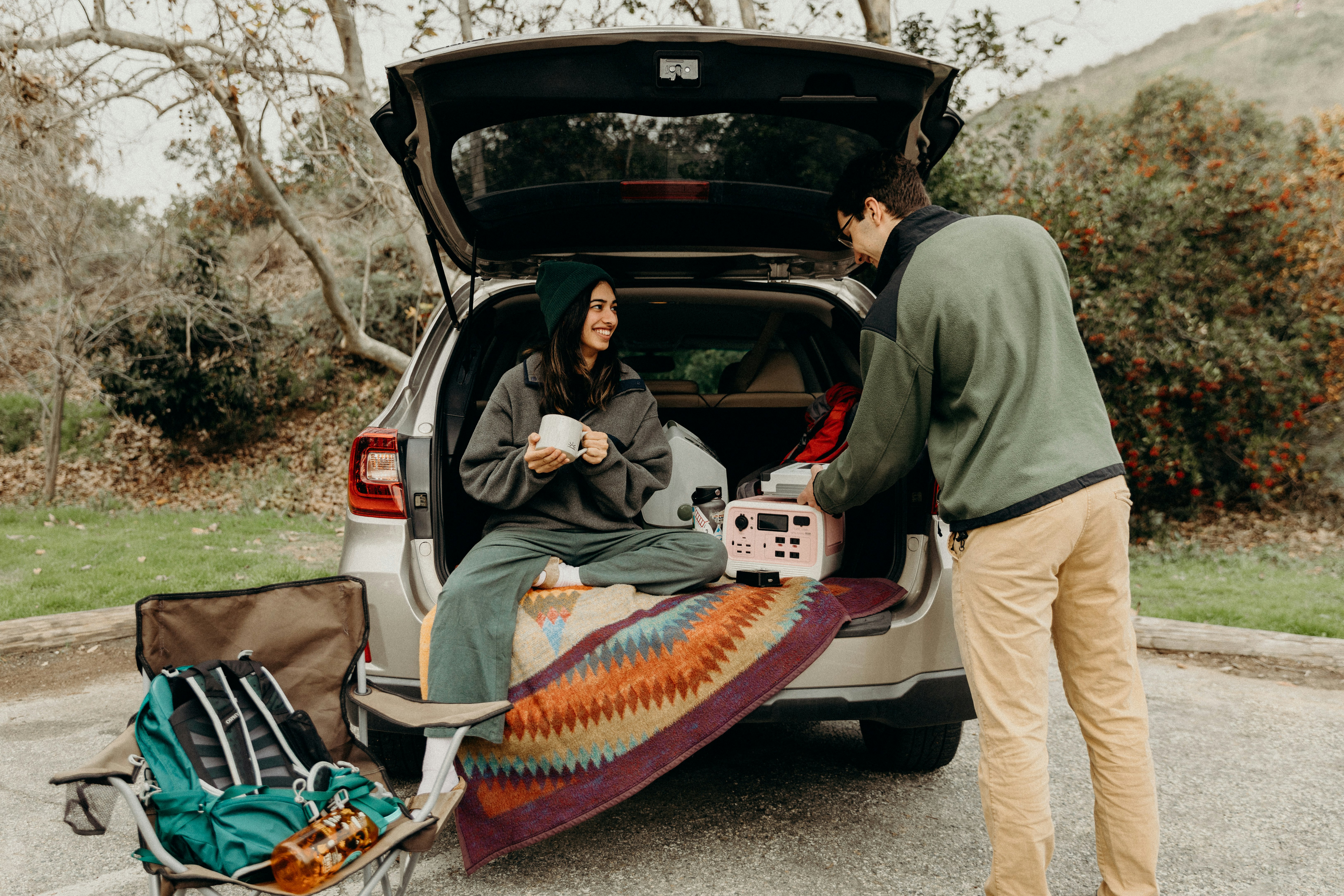 Two people prepare for outdoor activities at the open trunk of a car; one sits on a blanket with a mug, while the other loads a pet carrier | outdoorsurvivors com