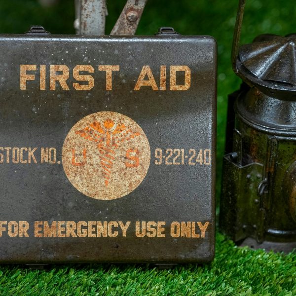 A metal first aid box labeled “For Emergency Use Only” sits on grass next to a vintage lantern. | outdoorsurvivors.com A metal first aid box labeled “For Emergency Use Only” sits on grass next to a vintage lantern | outdoorsurvivors com
