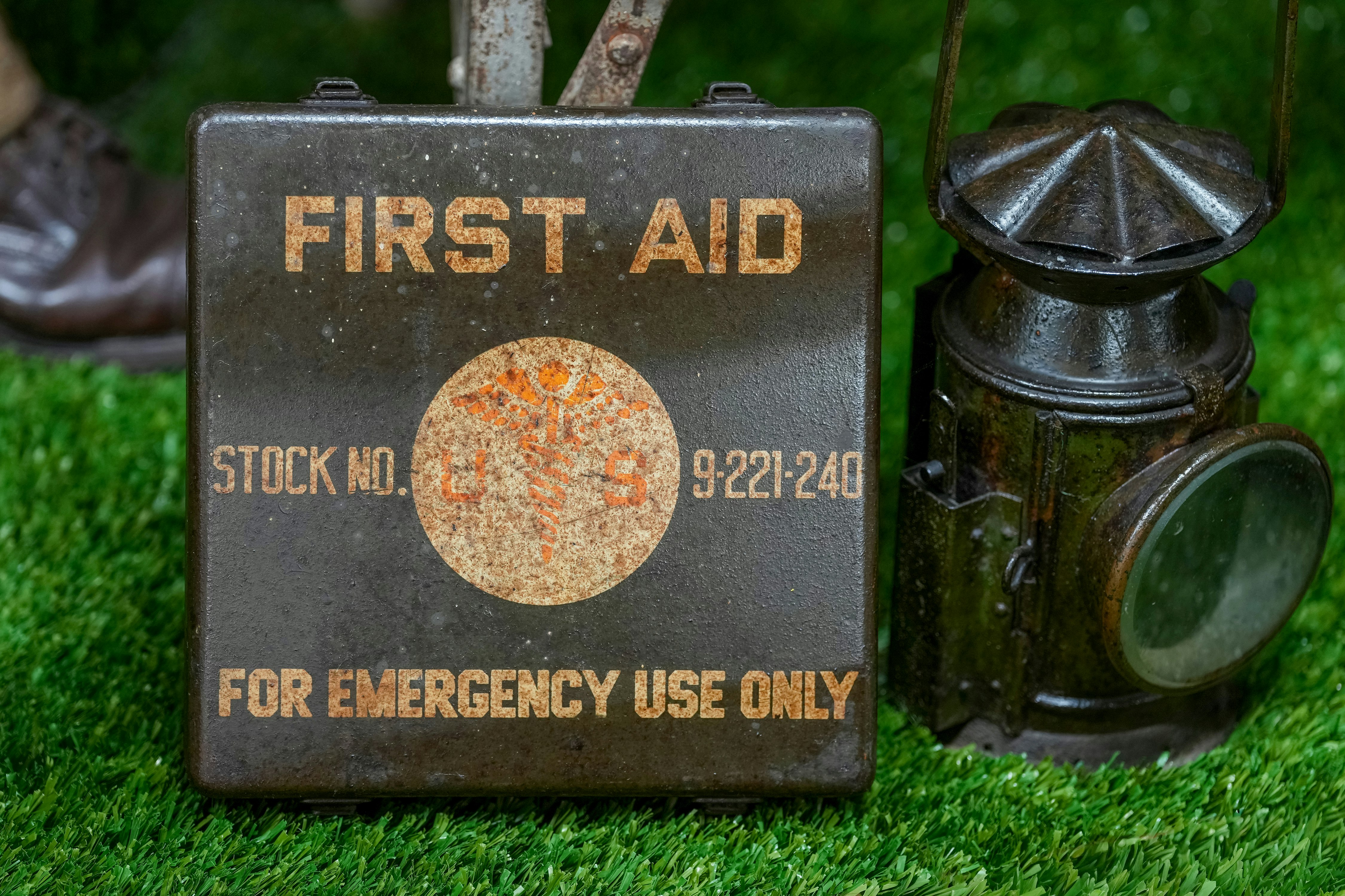 A metal first aid box labeled “For Emergency Use Only” sits on grass next to a vintage lantern | outdoorsurvivors com