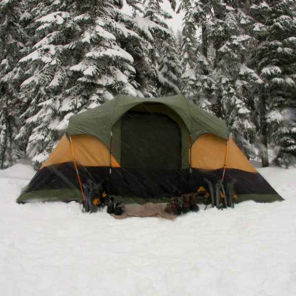 A green and yellow tent pitched in deep snow in front of snow-covered pine trees, with several pairs of boots lined up in front of the entrance. | outdoorsurvivors.com A green and yellow tent pitched in deep snow in front of snow covered pine trees, with several pairs of boots lined up in front of the entrance | outdoorsurvivors com