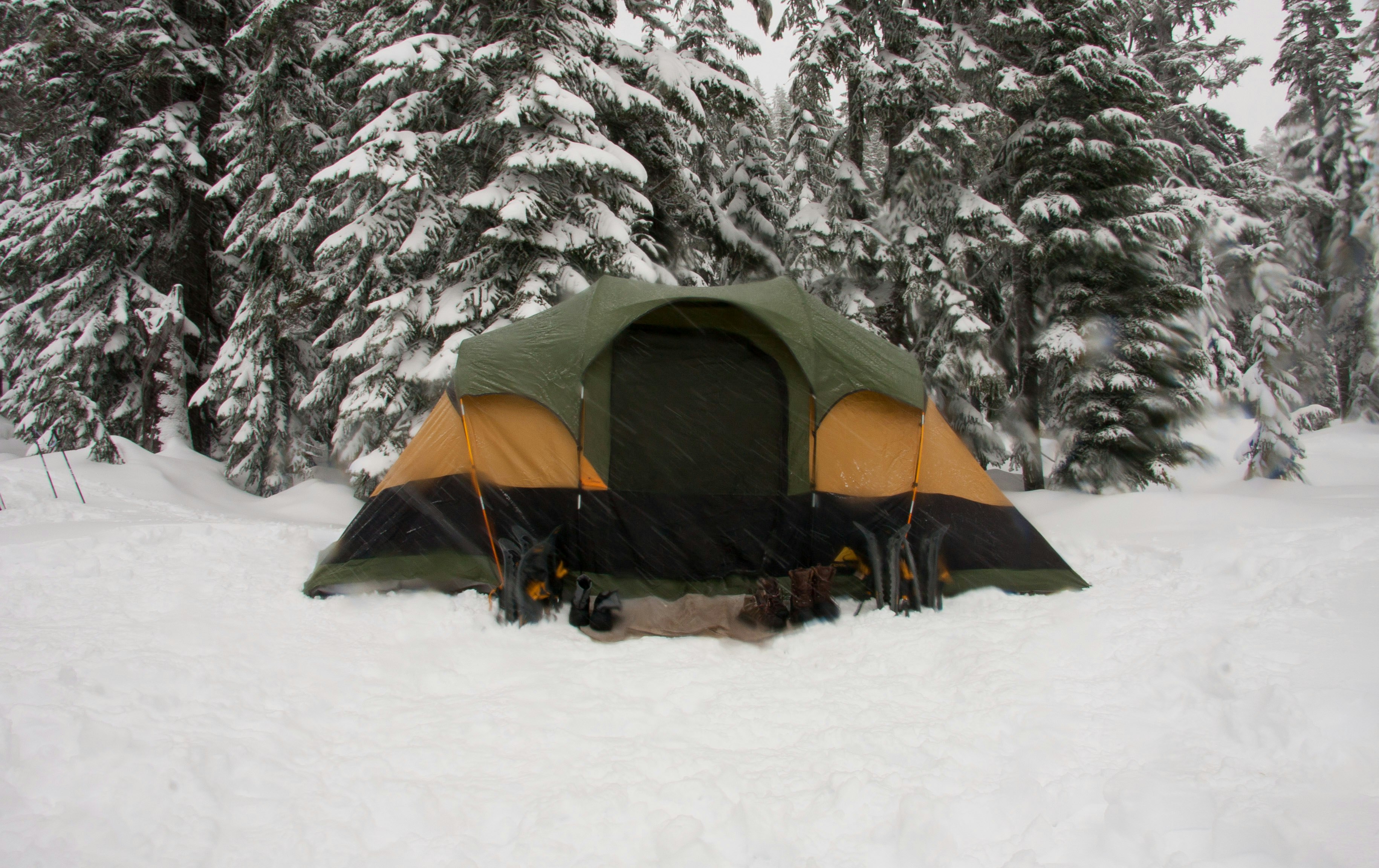A green and yellow tent pitched in deep snow in front of snow covered pine trees, with several pairs of boots lined up in front of the entrance | outdoorsurvivors com